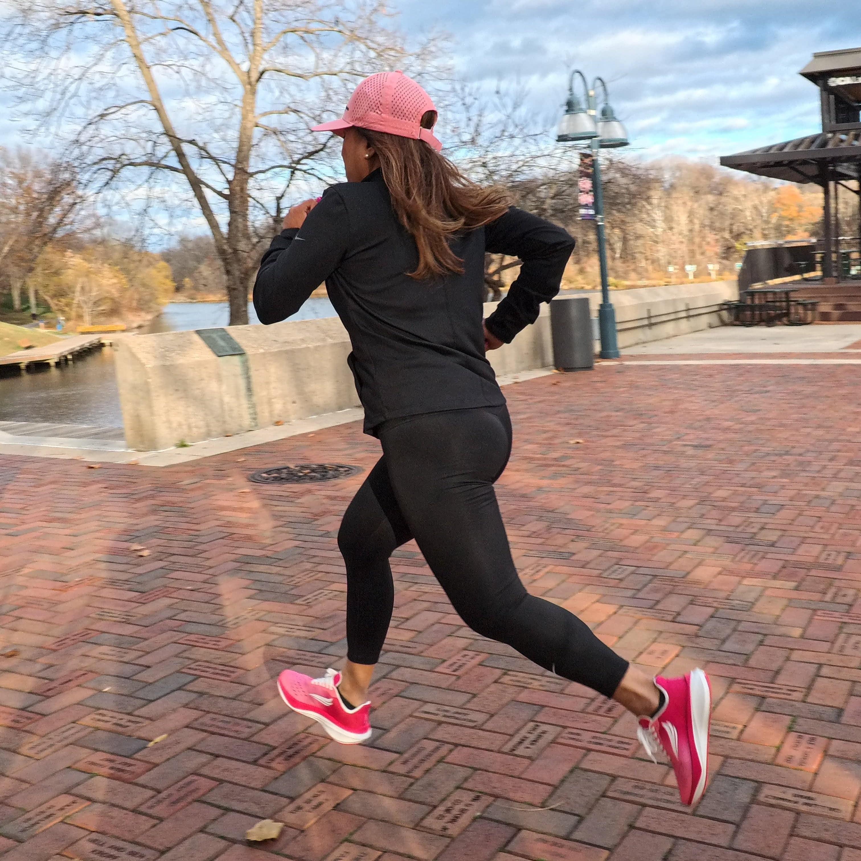 Woman running outdoors in pink athletic shoes, black leggings, and pink cap on brick path