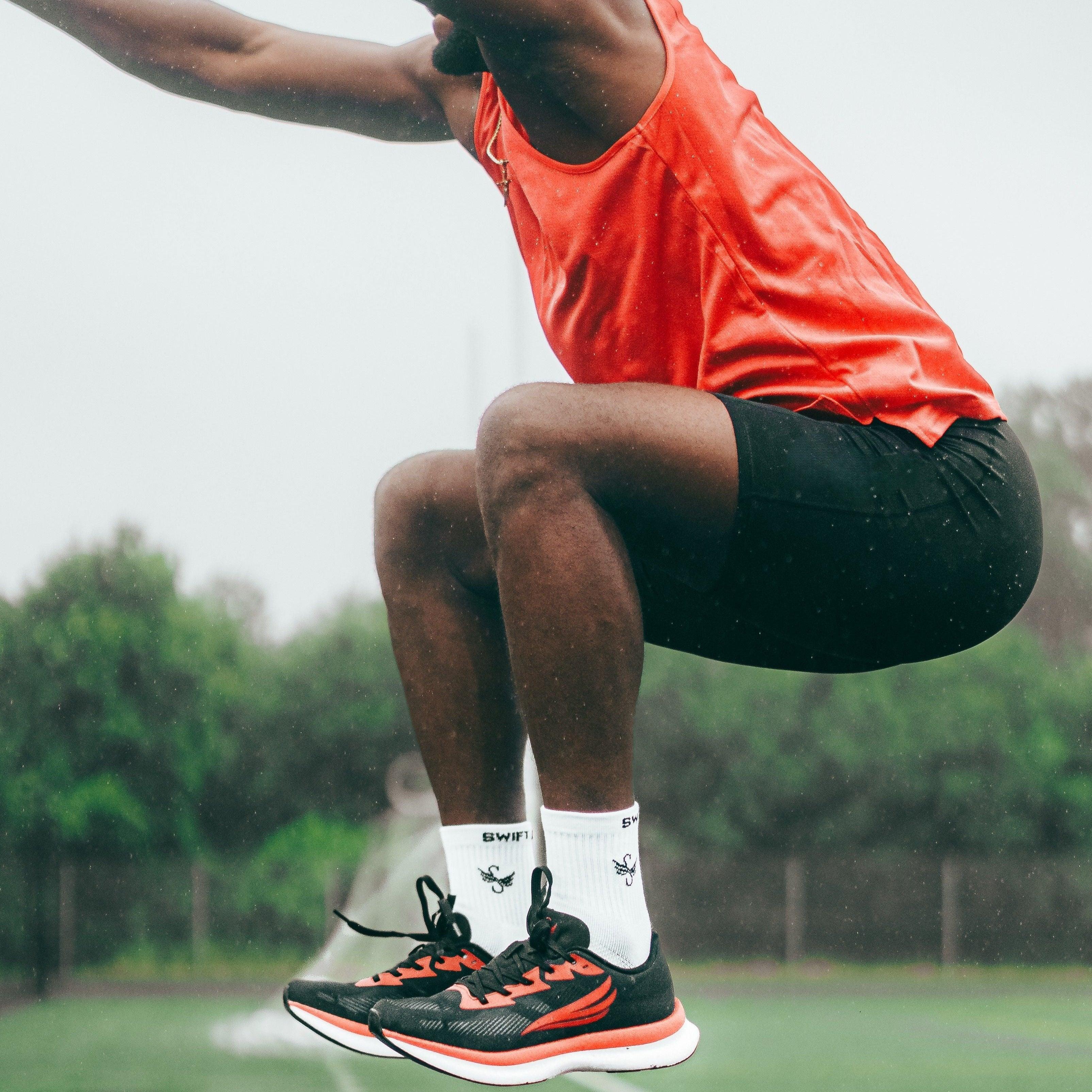 Athlete in red tank top and black shorts jumping outdoors in black and red athletic shoes