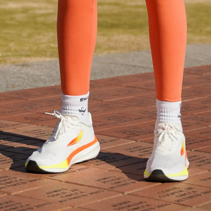 White athletic running shoes with orange and yellow accents worn outdoors on brick pavement