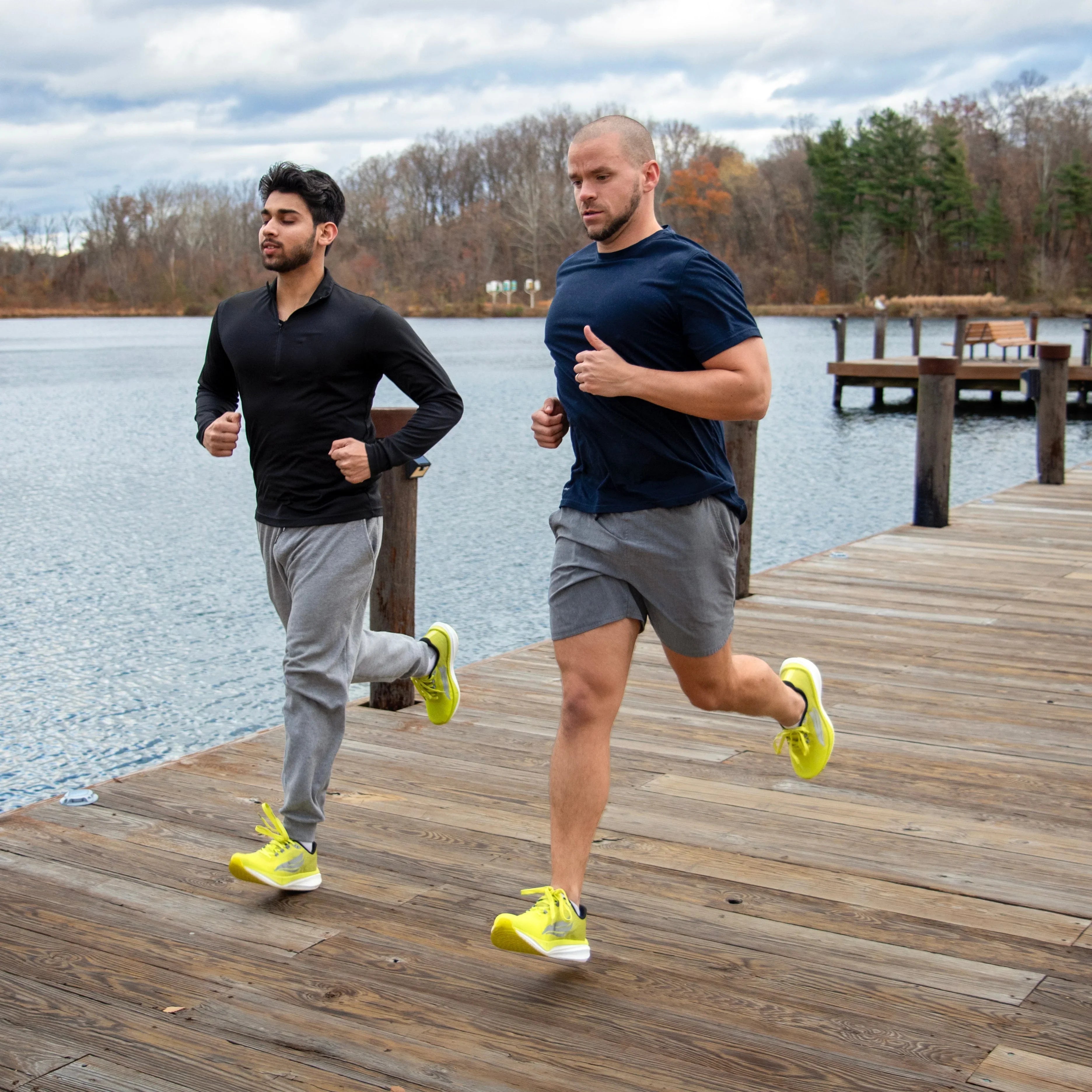 Two men running on a wooden dock by a lake wearing athletic shoes and workout clothing