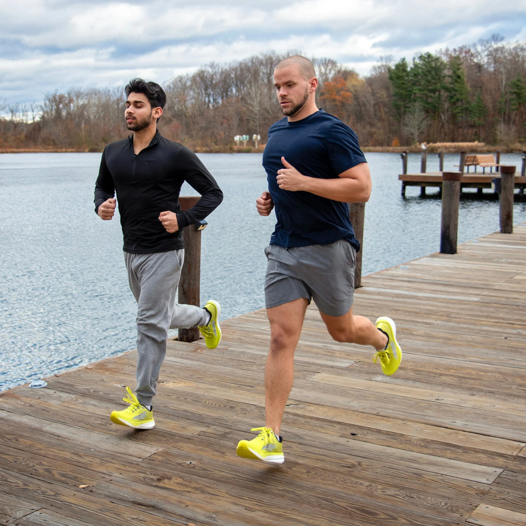 Two men running on a wooden dock by a lake wearing athletic shoes and workout clothing