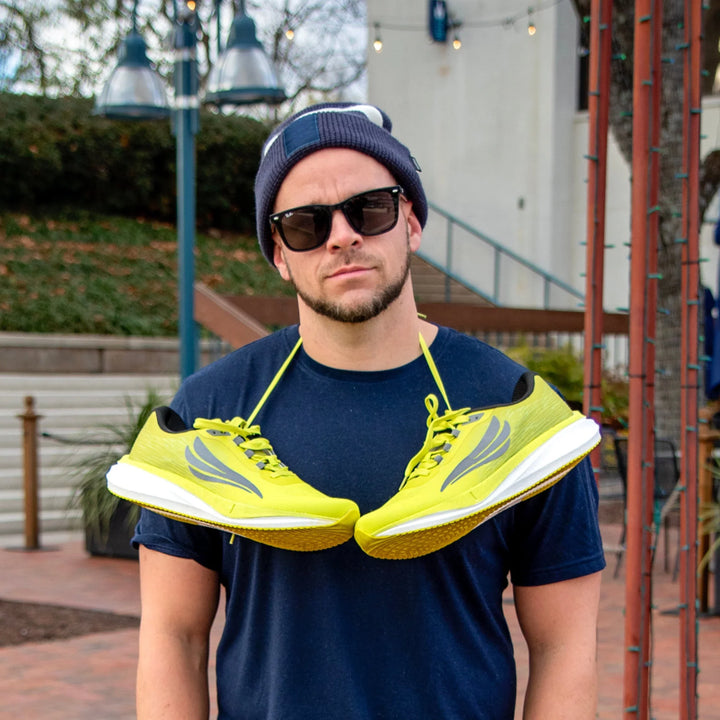 Man outdoors wearing sunglasses and a beanie with yellow running shoes around his neck