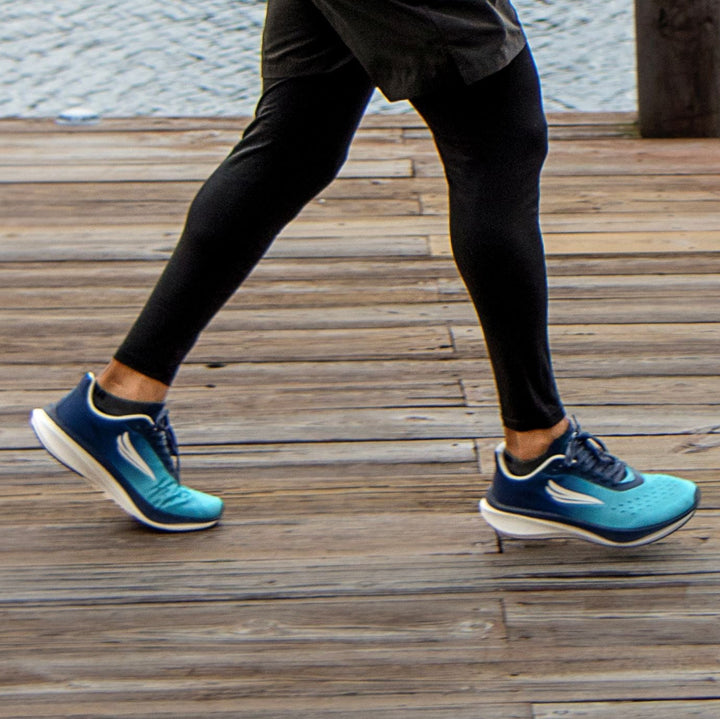 Person wearing blue running shoes and black leggings jogging on a wooden dock by water